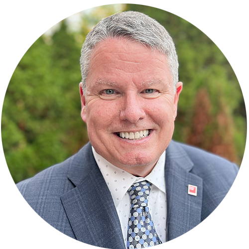 Kevin is in a blue suit as he smiles for a photo outside in front of green trees.