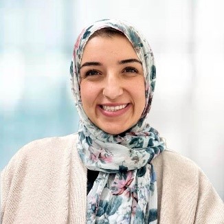 Nagham is smiling for a professional headshot. The image is brightly lit with a light blue and white background.