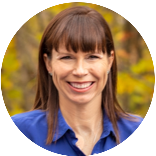 Noella is smiling for a headshot outside in front of a forest whose leaves are turning yellow in fall.