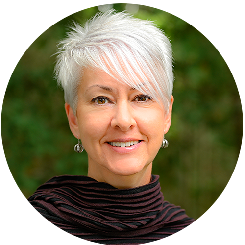 Jenn is smiling for a professional headshot, she'd standing outside on a sunny day in front of a green forest.